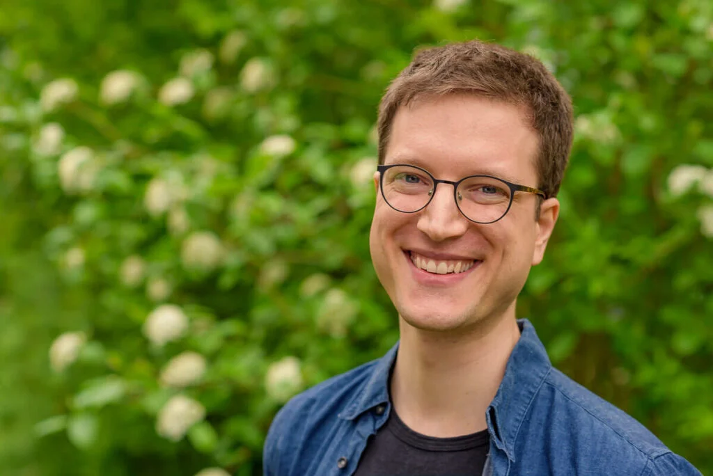 Porträt eines lächelnden Mannes mit Brille vor grünem, blühendem Hintergrund.