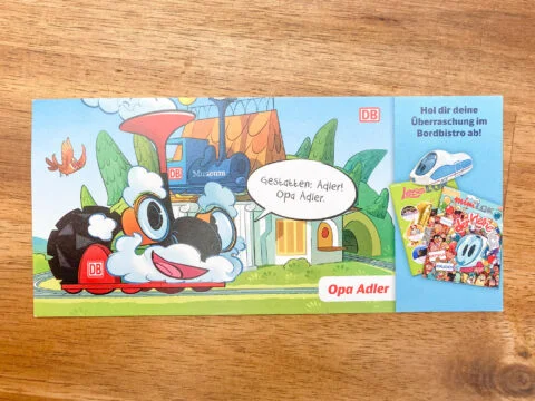 Illustration einer lachenden Dampflok mit Gesicht und Sprechblase, daneben zwei bunte Kinderzeitschriften und ein ICE-Zugmodell auf blauem Hintergrund.