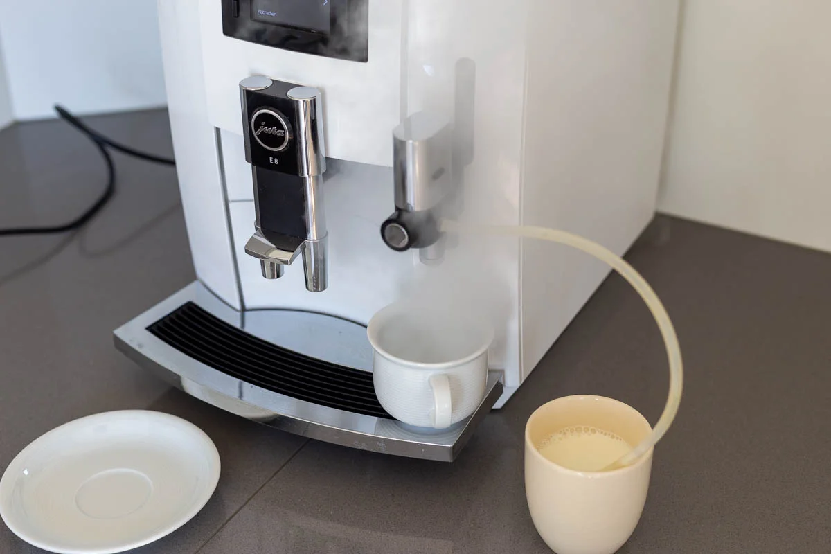 Weiße Jura-Kaffeemaschine mit dampfender Tasse und Milchschlauch, daneben eine Tasse mit Milch und eine Untertasse auf grauer Arbeitsfläche.