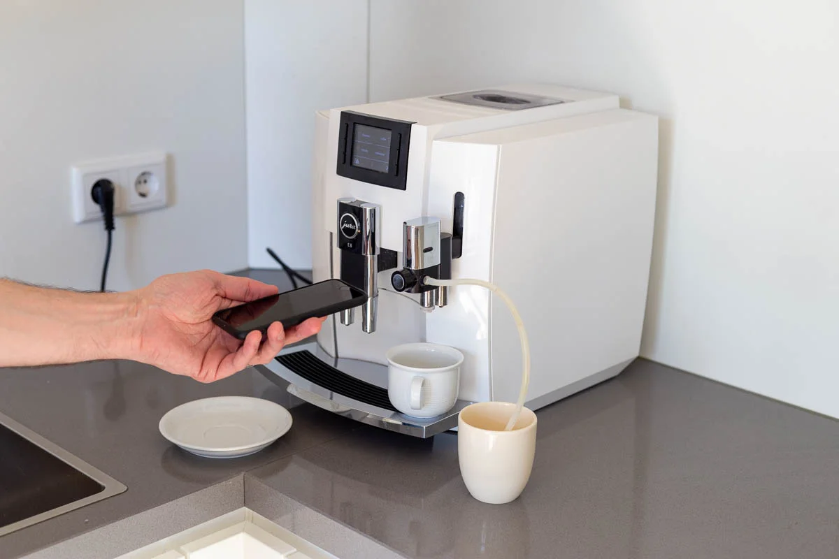 Eine Hand hält ein Smartphone vor eine weiße Kaffeemaschine auf einer Küchenarbeitsplatte mit zwei Tassen und einem Unterteller.