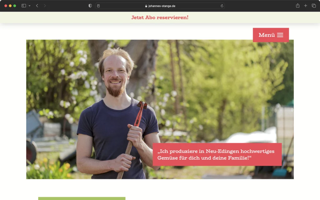 Mann mit Bart und T-Shirt hält eine Hacke in einem Garten mit grünen Bäumen und Pflanzen im Hintergrund.