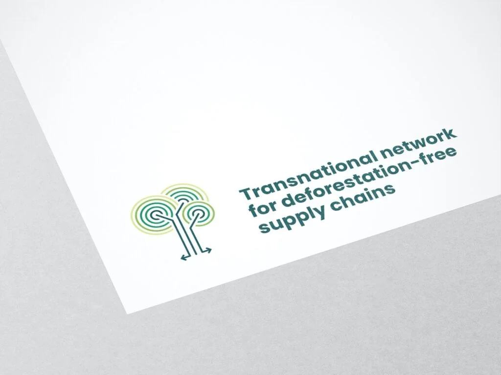 Logo mit stilisierten Bäumen und dem Text Transnational network for deforestation-free supply chains auf weißem Papier vor grauem Hintergrund.