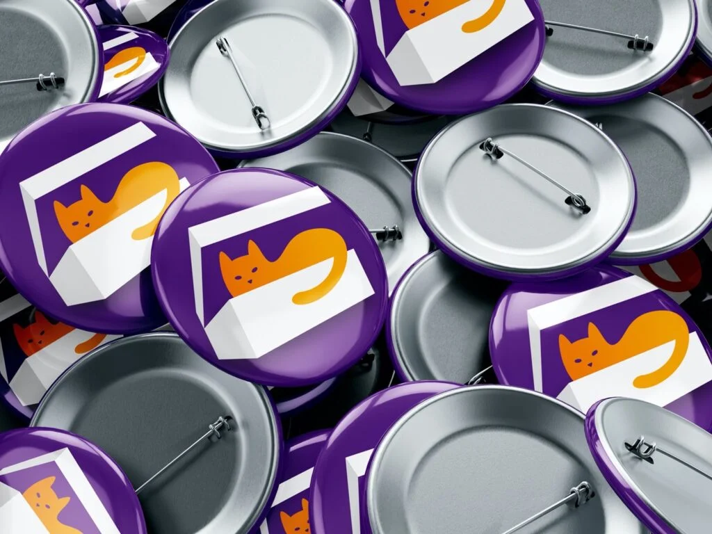 Viele lila Ansteckbuttons mit einer Grafik einer orangefarbenen Katze in einer weißen Box, einige Rückseiten mit Sicherheitsnadeln sichtbar.