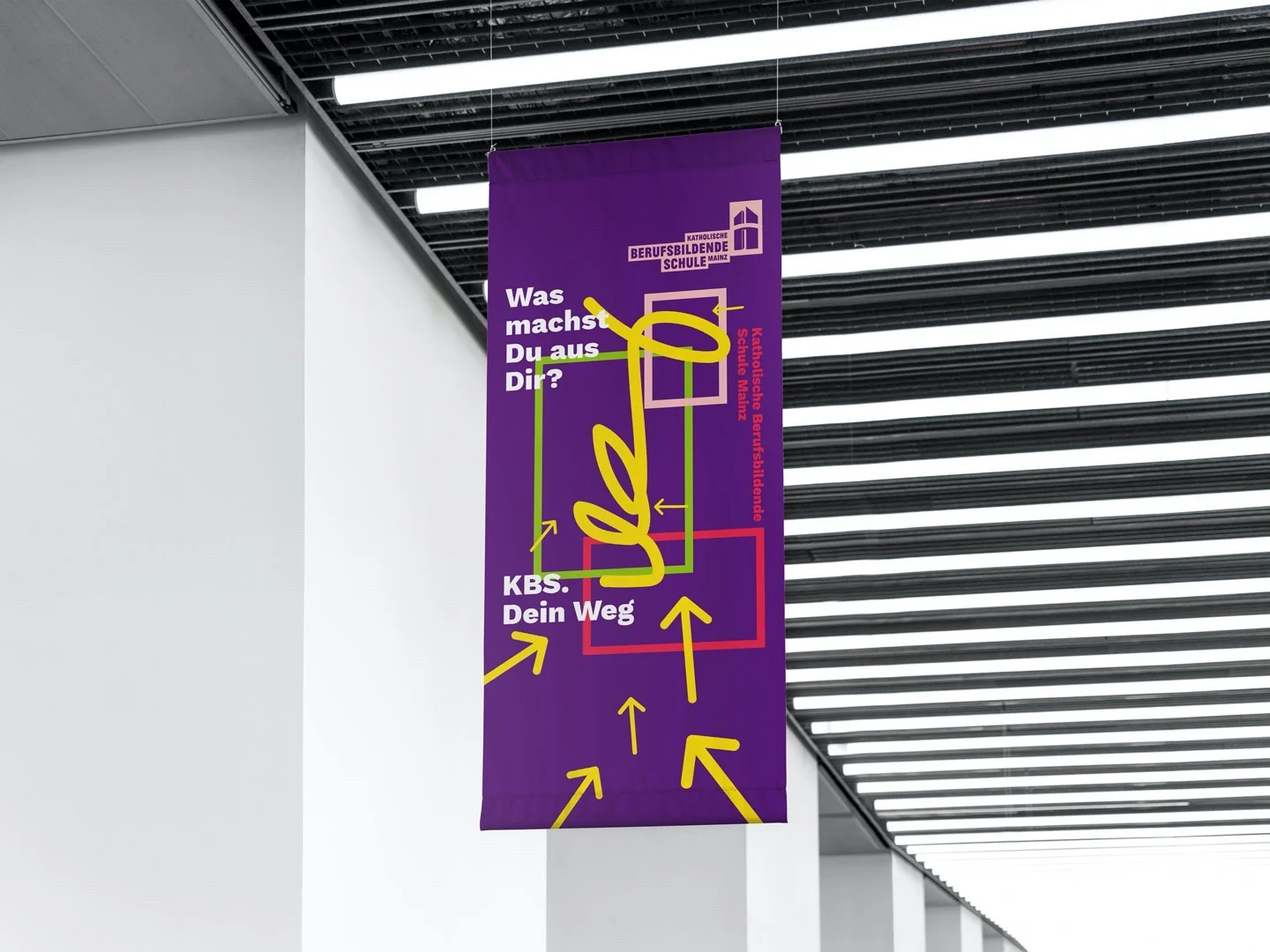 Ein lila Banner mit gelben Pfeilen und Text zur Katholischen Berufsbildenden Schule Mainz hängt in einem modernen Gebäude.