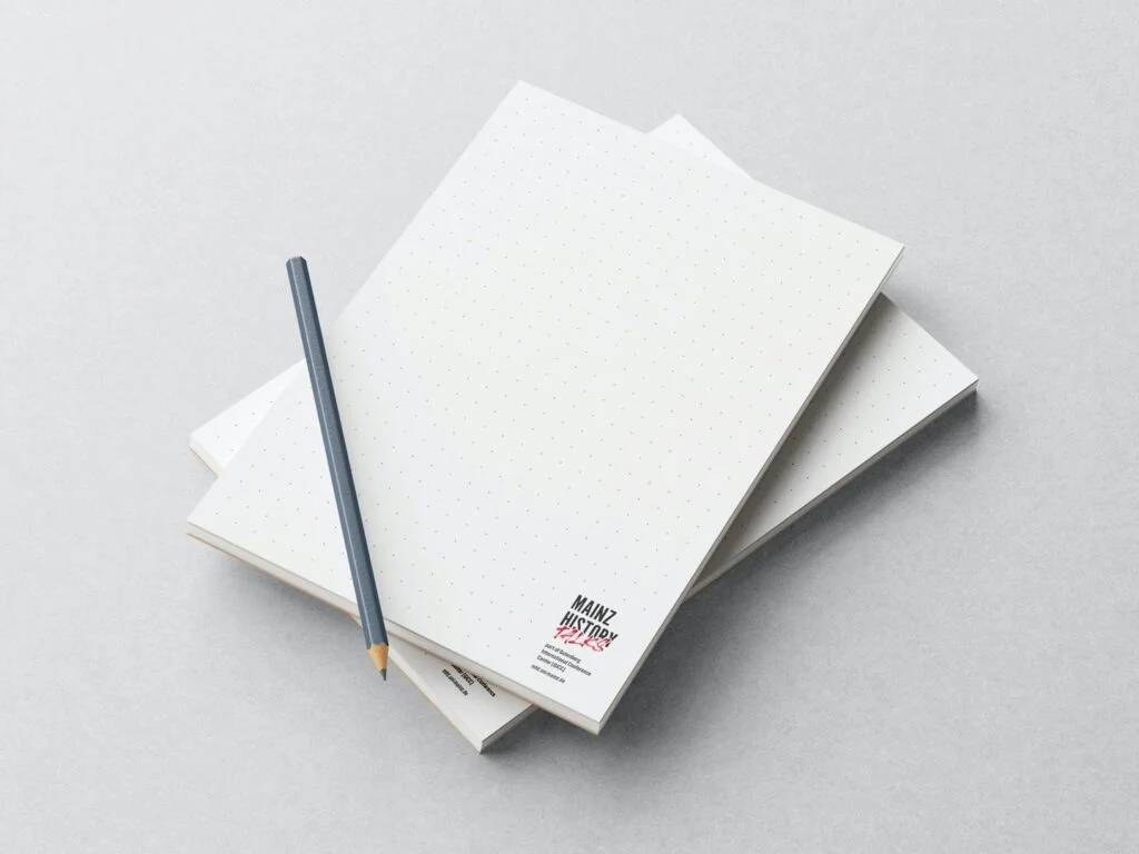 Zwei punktkarierte Notizbücher liegen übereinander auf einem hellen Untergrund, darauf liegt ein grauer Bleistift.