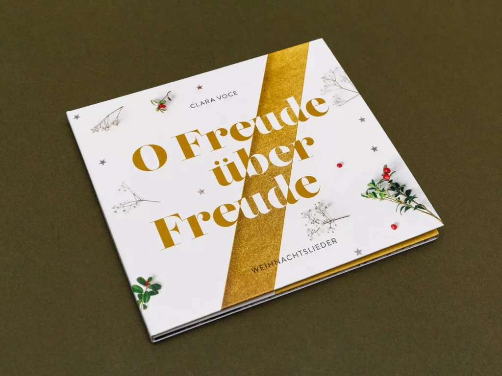 CD-Hülle mit dem Titel O Freude über Freude von Clara Voce und dem Zusatz Weihnachtslieder, dekoriert mit kleinen Zweigen und roten Beeren.