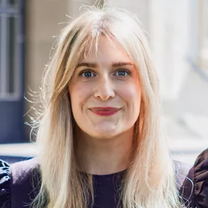 Porträt einer lächelnden Frau mit langen blonden Haaren vor unscharfem hellem Hintergrund.