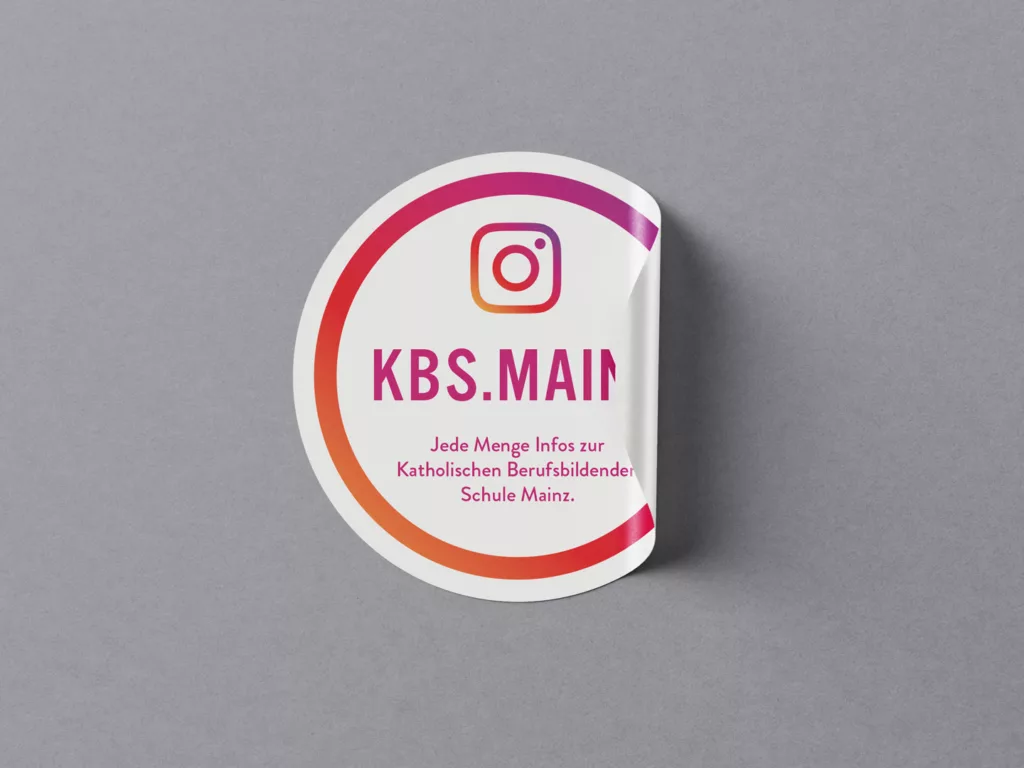 Aufkleber mit Instagram-Logo und Text KBS.MAIN sowie Hinweis auf Infos zur Katholischen Berufsbildenden Schule Mainz auf grauem Hintergrund.
