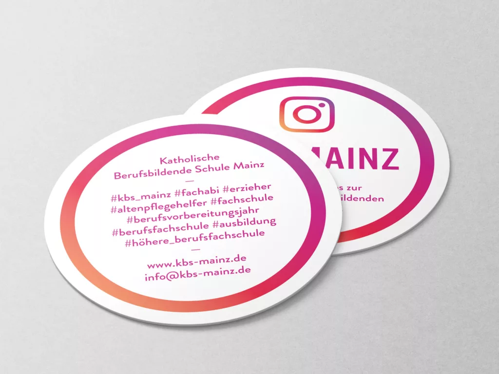 Zwei runde Aufkleber mit Instagram-Design und Texten zur Katholischen Berufsbildenden Schule Mainz, Hashtags und Kontaktinformationen.
