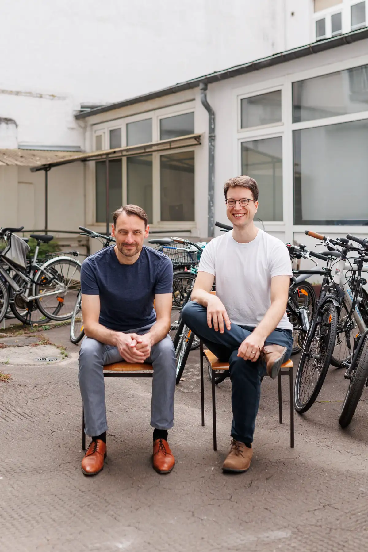 Zwei Männer sitzen auf Stühlen im Hof, umgeben von Fahrrädern, der linke Mann trägt ein blaues Shirt, der rechte ein weißes.
