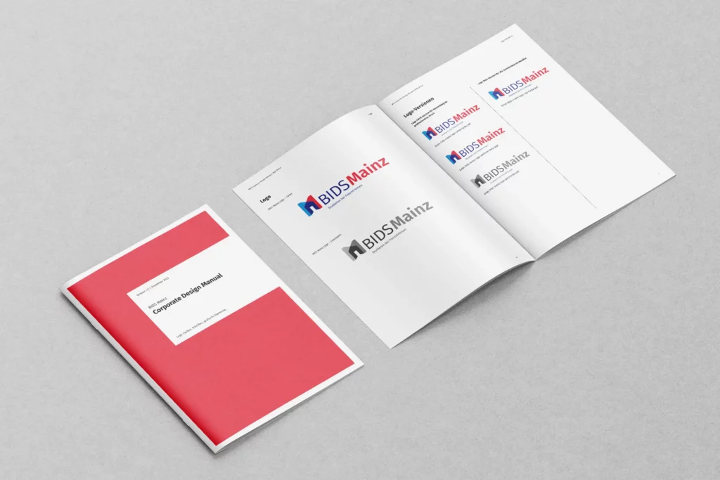 Zwei Corporate Design Manuals von BIDS Mainz liegen auf grauem Untergrund, eines geschlossen mit rotem Cover, das andere geöffnet mit verschiedenen Logo-Versionen.